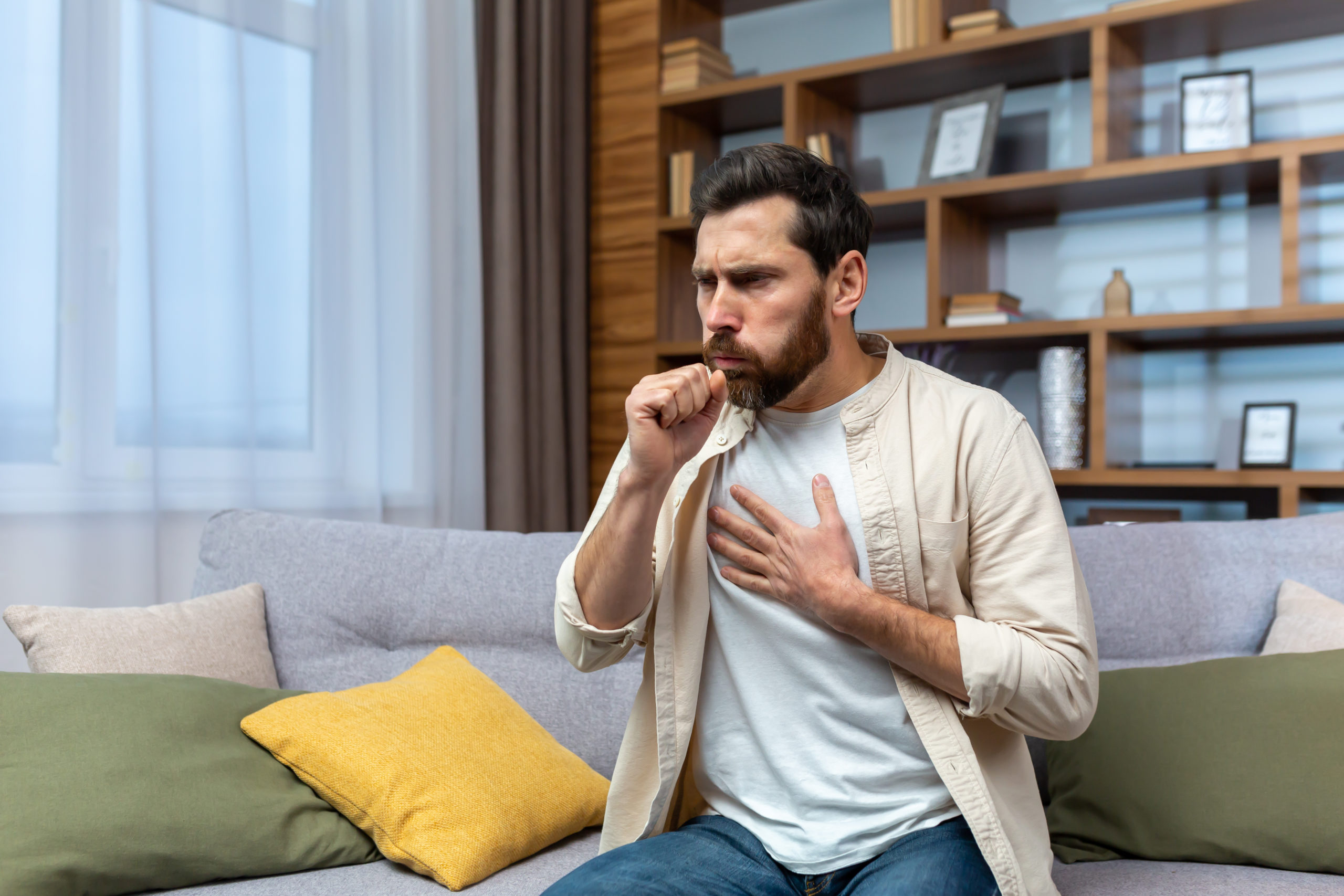 Sick mature man alone at home sitting on sofa coughing holding hands to chest in living room.